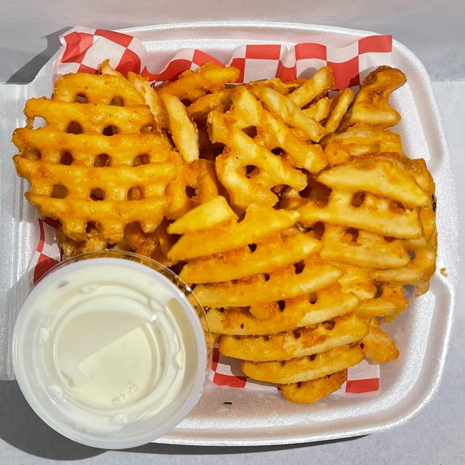 Golden waffle fries with seasoning and a side of white dipping sauce in a styrofoam container.