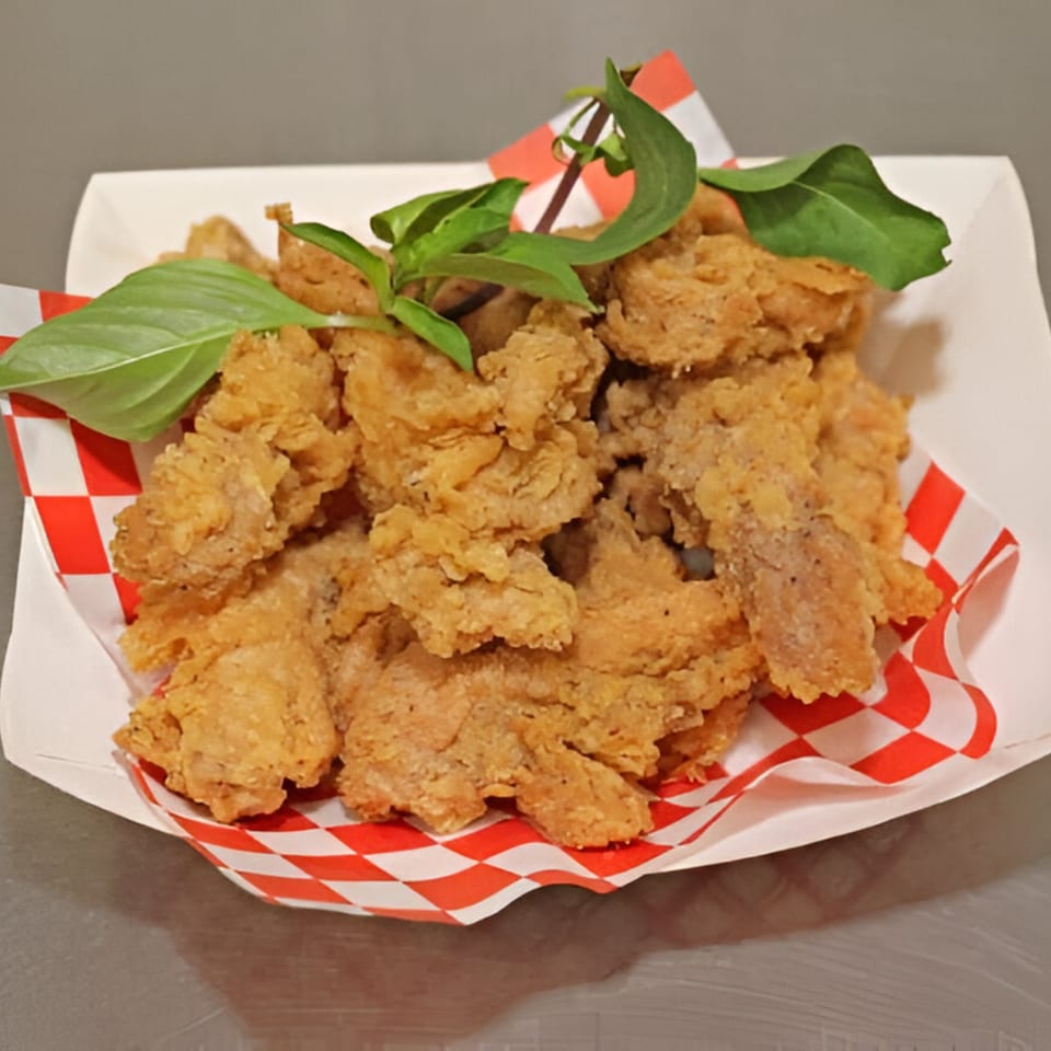 Crispy fried chicken pieces with fresh basil in a white paper tray with red and white checkered lining.