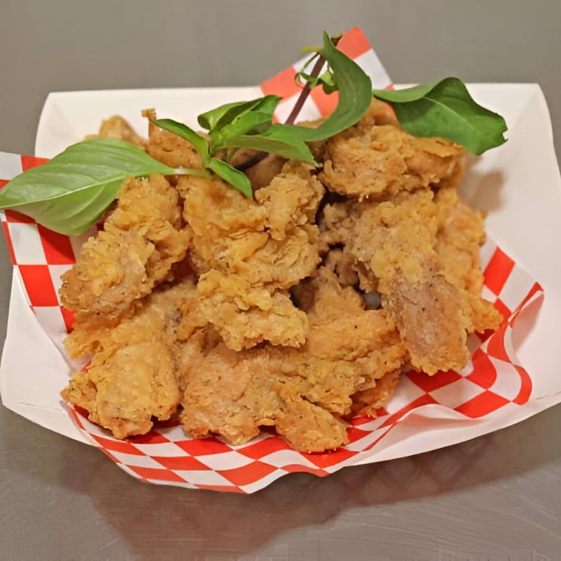 Crispy fried chicken pieces with fresh basil in a white paper tray with red and white checkered lining.
