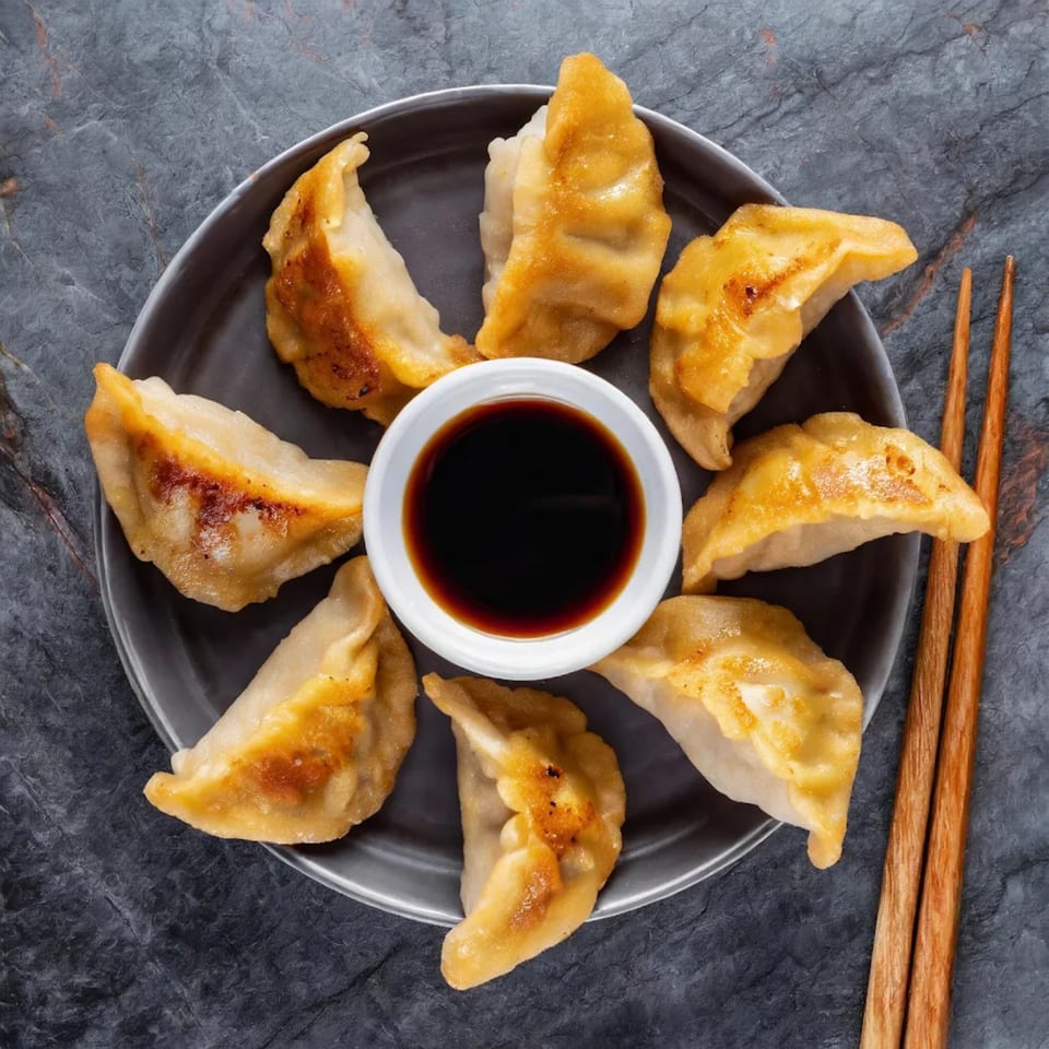 Golden-brown potstickers arranged around a white bowl of soy sauce, with chopsticks on a dark surface.